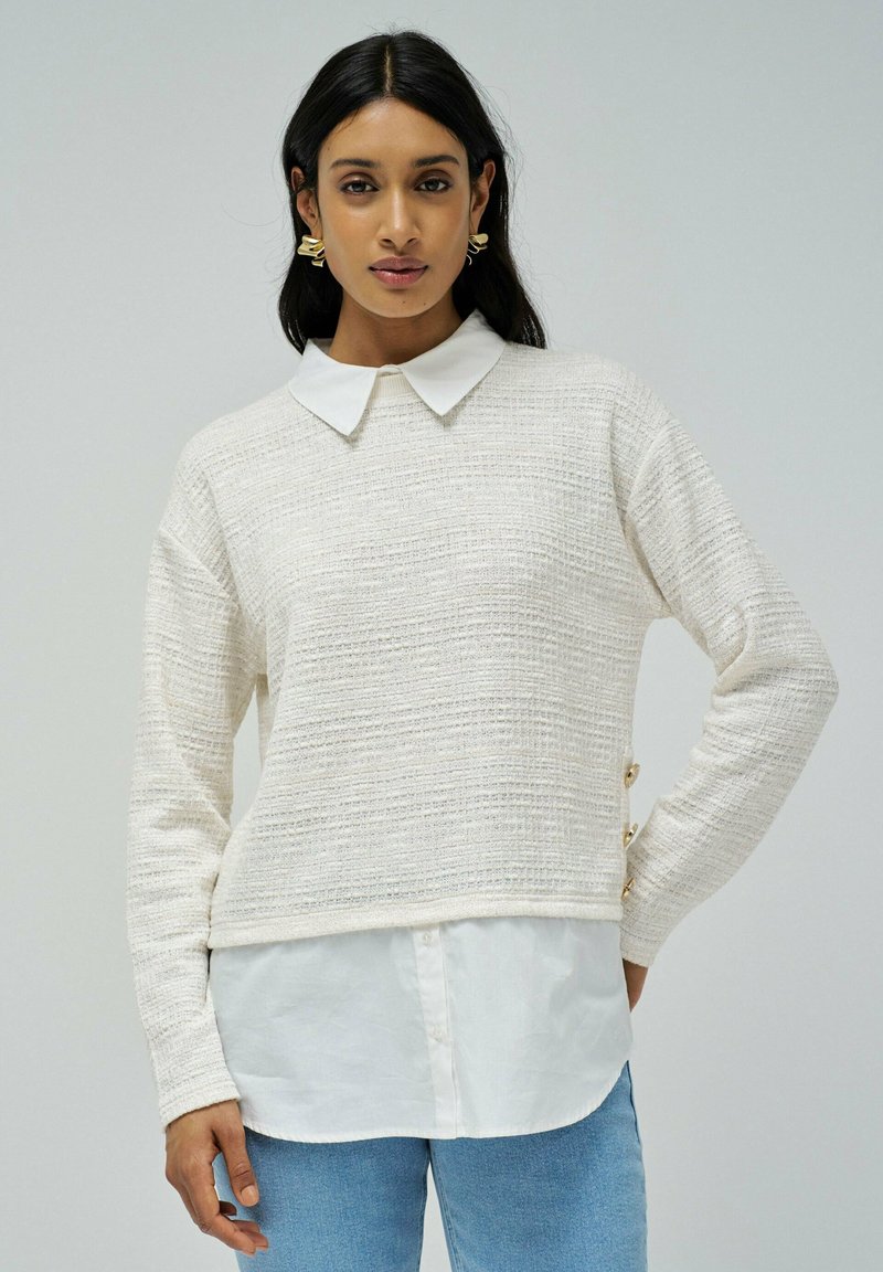 Pull en maille beige clair avec un motif texturé, manches longues, et une chemise blanche à col en dessous. Présente des boutons décoratifs sur le côté.