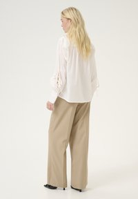Blusa bianca con maniche a palloncino e dettaglio a fiocco, abbinata a pantaloni beige larghi e scarpe slip-on nere, vista da dietro.