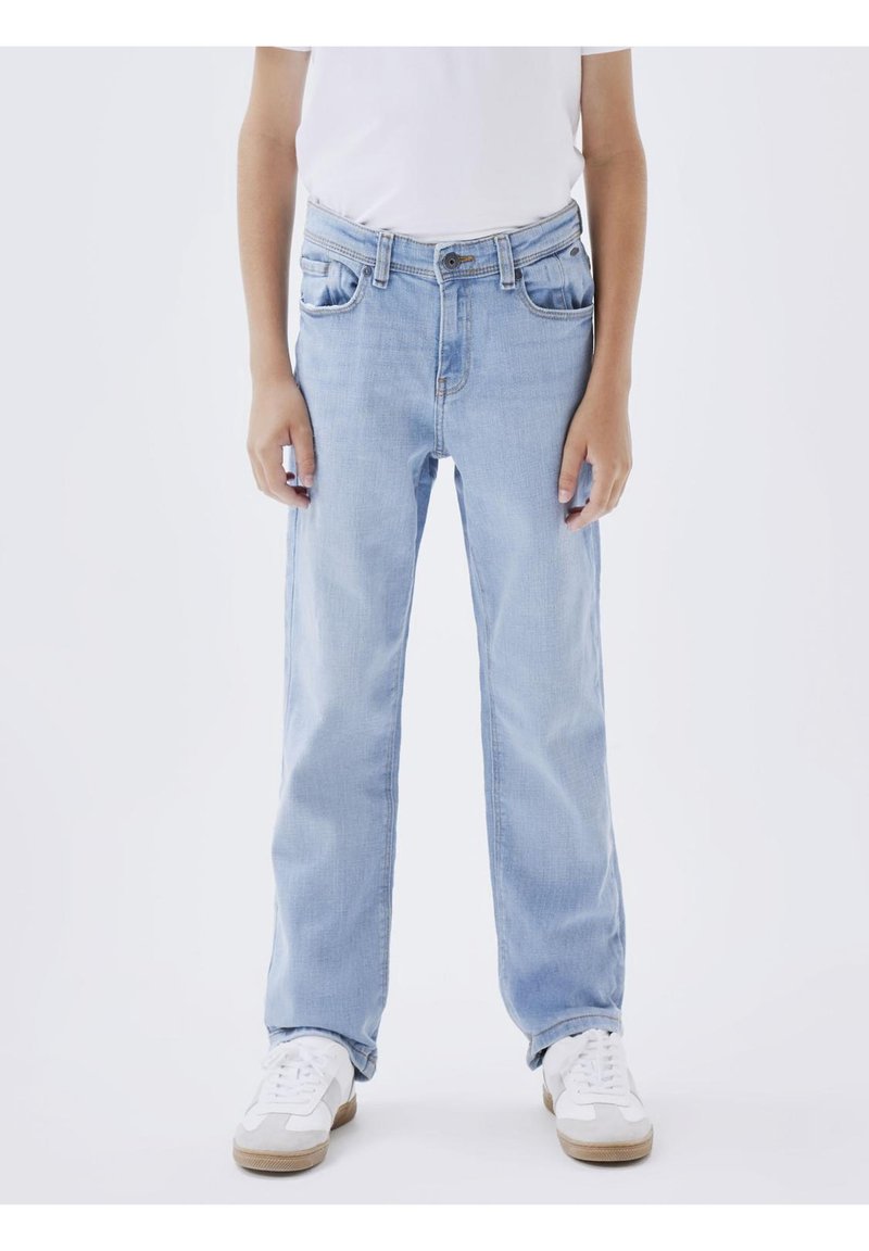 Personne portant un jean droit bleu clair, des baskets blanches et une chemise blanche, debout devant un fond blanc uni.
