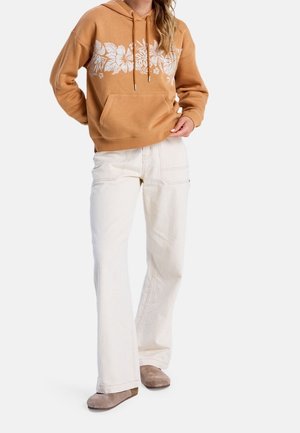 Person iført en brun hættetrøje med hvidt blomsterprint, off-white vidde bukser og taupe slip-on sko, stående mod en hvid baggrund.