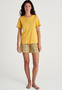 Geel katoenen t-shirt met korte mouwen, gecombineerd met een gekleed short met een bloemenpatroon in gele, witte en grijze tinten.