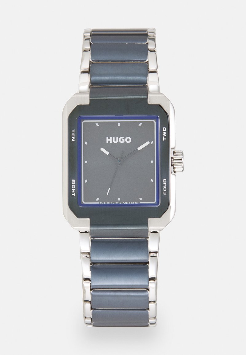 Kona hõbedane kell, millel on tumehall sihverplaat, sinised aktsendid ja roostevabast terasest rihm. Veekindel kuni 50 meetrit.