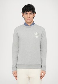 Szara bluza z długimi rękawami, okrągłym dekoltem i ści�ągaczami przy rękawach. Na lewej piersi znajduje się biały logotyp, w zestawieniu z jasnymi spodniami.