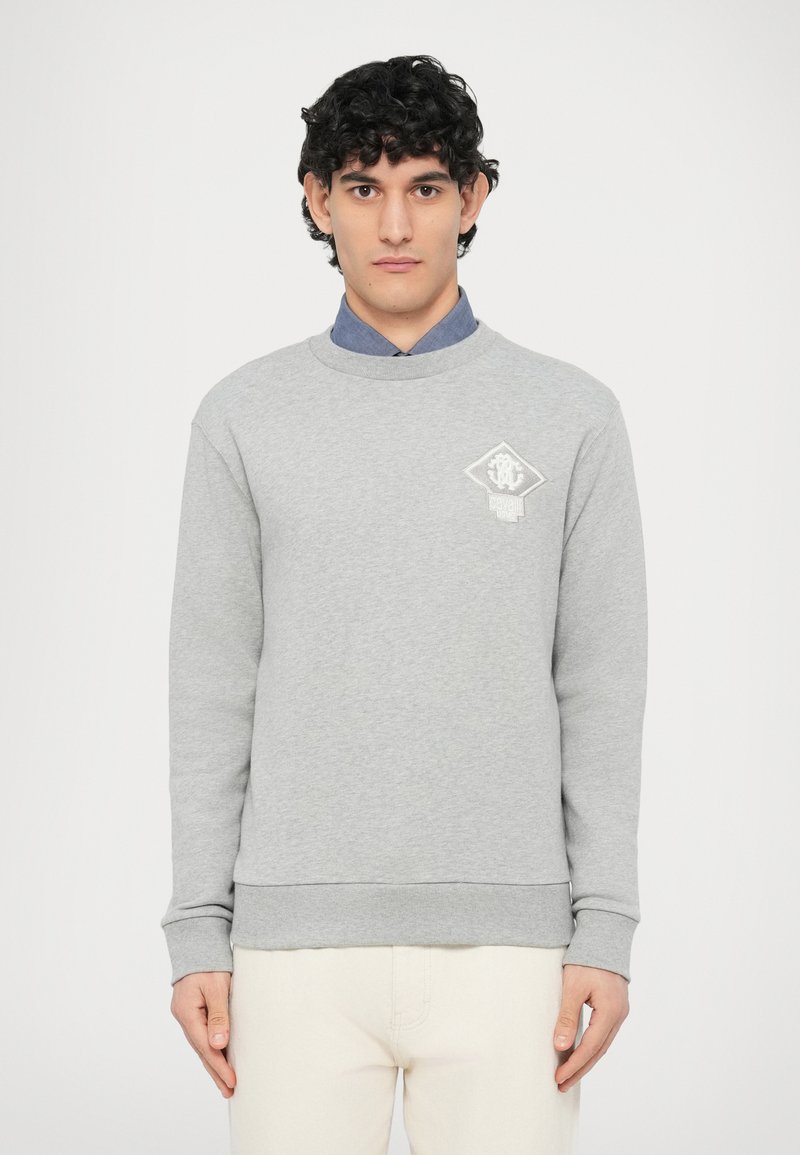 Szara bluza z długimi rękawami, okrągłym dekoltem i ściągaczami przy rękawach. Na lewej piersi znajduje się biały logotyp, w zestawieniu z jasnymi spodniami.