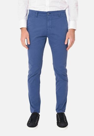 Uomo che indossa pantaloni blu slim-fit, camicia bianca abbottonata e sneakers nere con suola bianca, in piedi davanti a uno sfondo uniforme.
