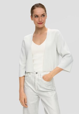 Weißer, kurz geschnittener Cardigan mit Dreiviertelärmeln, strukturiertem Stoff, offener Vorderseite, kombiniert mit einem weißen V-Ausschnitt-Oberteil und hellen Hosen.