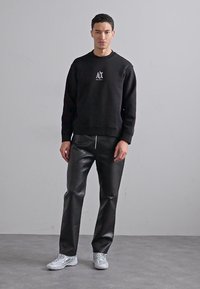 Black sweatshirt with a subtle logo, paired with shiny black faux leather pants. Model wears light sneakers, standing against a neutral background.