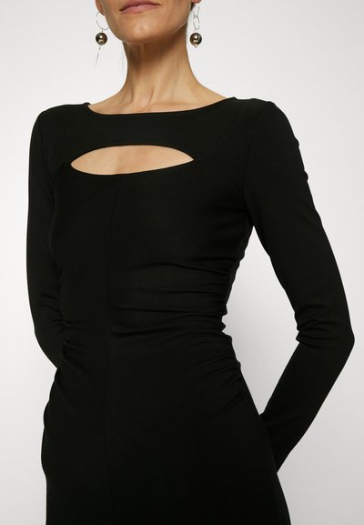 Μαύρο φόρεμα με μακρύ μανίκι, με κοψίματα σε σχήμα κλειδί στη λαιμόκοψη. Μαλακό, υφασματώδες υλικό με πτυχώσεις στα πλάγια.