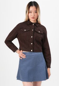 Kvinne iført en brun leopardmønstret denimjakke med sølvknapper og et blått cordfløyelskjørt, poserer med en hånd på hoften.