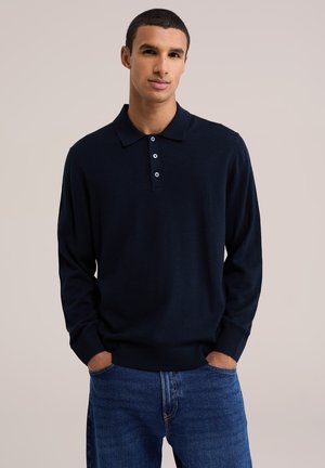 Giovane uomo con capelli corti che indossa una polo a maniche lunghe scura e jeans blu, in piedi con le mani nelle tasche davanti a uno sfondo uniforme.