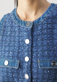 Gros plan sur une veste en tweed bleu avec de petites paillettes, des boutons argentés et un bord en denim autour du col et des poches sur le haut du torse d'une personne.