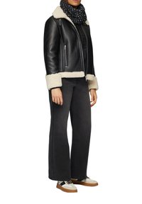 Veste en cuir noir avec col en fausse fourrure, associée à un pantalon en denim foncé évasé et une écharpe noire à motifs. Les chaussures sont blanches avec des accents bruns.