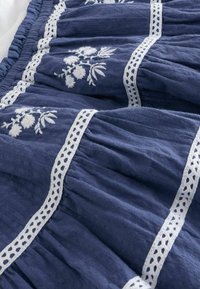 Marinblått tyg med vit blommig broderi och spetskant. Materialet ser mjukt ut med samlade veck som skapar en strukturerad look.