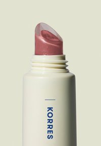 Bâton de cosmétique Korres, contenant blanc avec texte bleu, doté d'un cap transparent et d'un embout rose scintillant. Design lisse et arrondi.