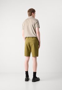 Beige polo skjorte med korte ermer, parret med olivengrønne shorts i knæ længde. Sorte sko og sokker fuldender outfittet. Ensfarvet baggrund.