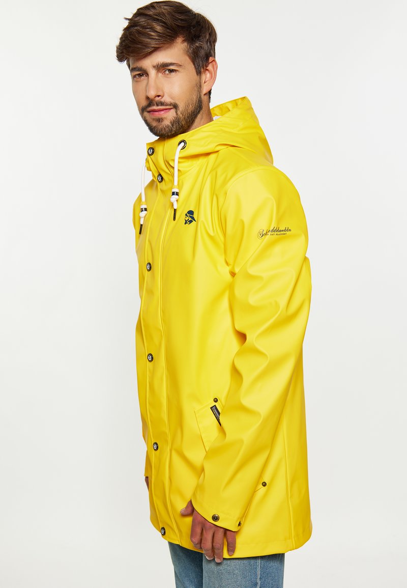 Geel waterdichte regenjas met een capuchon, drukknoopsluiting en een voorzak. Heeft trekkoorden en een logo op de borst. Glad van textuur.