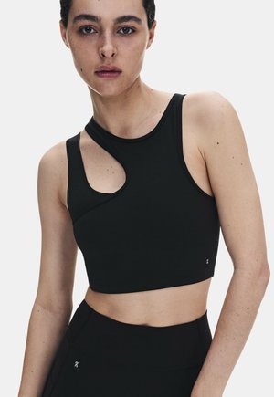 Jeune femme aux cheveux courts et foncés portant un crop top de sport asymétrique noir et des leggings noirs taille haute sur un fond uni.