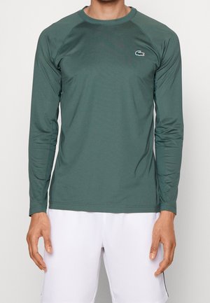 Homme portant un maillot de sport à manches longues vert foncé avec un petit logo de crocodile sur la poitrine et un short blanc, debout devant un fond uni.