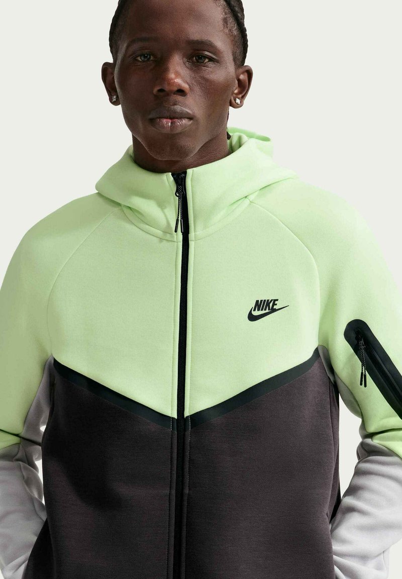 Mand iført lysegrøn, sort og hvid Nike lynlås-trøje med høj krave og lynlåslomme på venstre ærme.