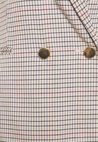 Blazer à carreaux blanc et rose avec des lignes noires et rouges, doté de deux grands boutons ronds métalliques près de la taille et d'une poche fendue horizontale.