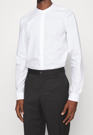 Man draagt een effen wit overhemd met lange mouwen en verborgen knoopsluiting, ingestopt in een donkere broek met hoge taille.