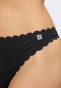 Juodas bikini apatinių dalis su išpjauta banguota kraštine. Juosmuo dekoruotas mažuoju baltu „HUGO“ logotipu. Lygi audinio tekstūra.
