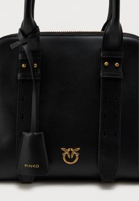 Μαύρη δερμάτινη τσάντα με χρυσές λεπτομέρειες. Περιλαμβάνει ραμμένες λαβές, ένα διακοσμητικό έμβλημα και κλείσιμο με φερμουάρ. Ομαλή υφή σε όλη την επιφάνεια.