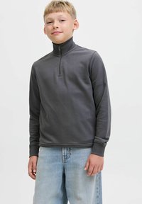 Garçon avec des cheveux blonds courts, portant un pull à col zippé gris foncé et un jean bleu clair, se tenant devant un fond blanc uni.