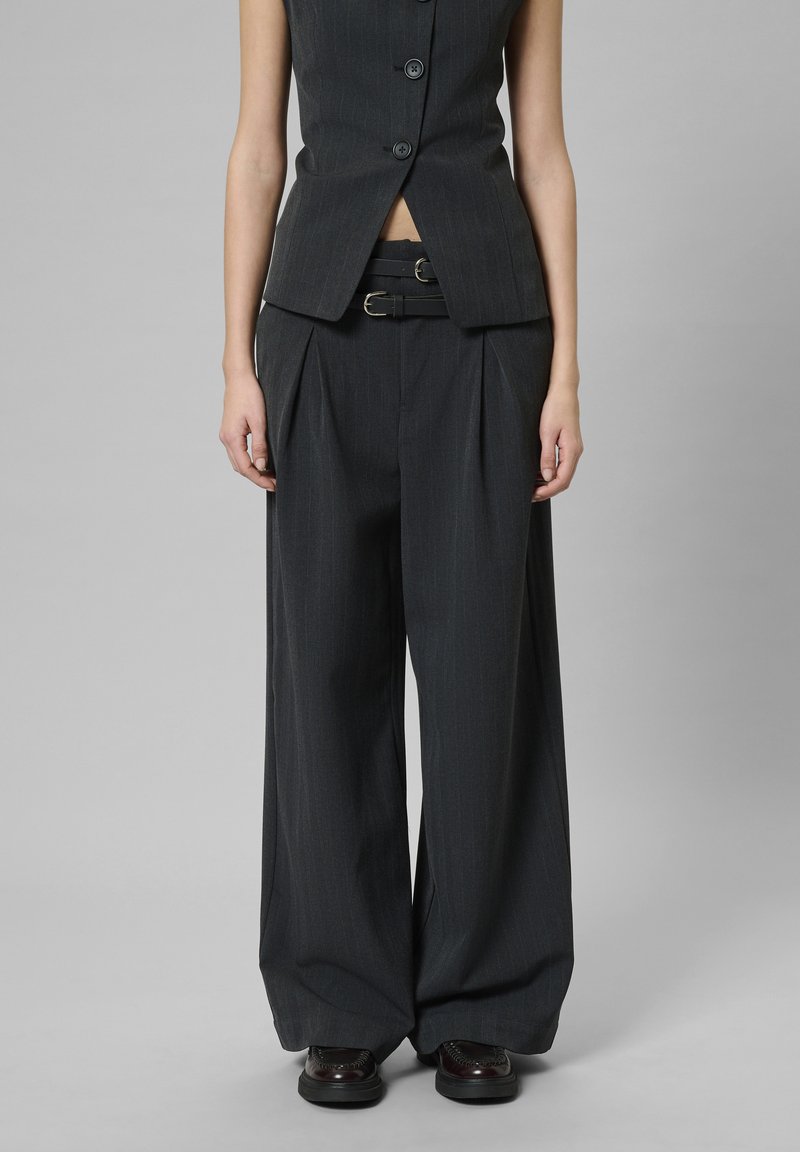 Fekete csíkos nadrág magas derékkal, redőkkel és hozzáillő övvel. Egy testhezálló, ujjatlan fekete mellénnyel viselve.