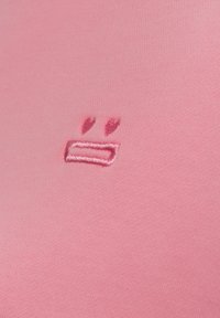 Rosa Stoff mit einem kleinen, gestickten neutralen Gesichtssymbol, bestehend aus zwei vertikalen Strichen über einem horizontalen rechteckigen Mund.