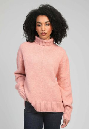 Femme aux cheveux bouclés portant un pull à col roulé rose et un jean foncé, debout les mains dans les poches devant un fond uni.