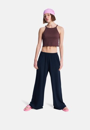 Bruin cropped tanktop met ruwe zoom, donkerblauwe brede broek en roze teenslippers. Accessoires omvatten een roze bandana en een kralenketting.