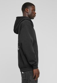 Sudadera negra con bolsillo delantero, corte relajado, puños acanalados y logotipo detallado en la parte posterior. Tejido suave, estilo casual, vista lateral.