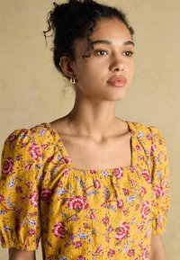 Kollane lilleline kleit puffed varrukatega ja ruudukujulise dekolteega. Omab punaseid ja siniseid lillemustreid tekstuurilisel kangal, millel on õrnad augud.