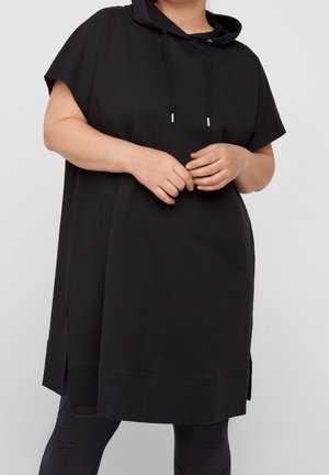 Osoba oblečená v volných černých šatech s kapucí a krátkými rukávy přes černé legíny, stojící s rukama sepnutýma před sebou.