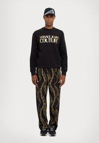 Black sweatshirt with gold "VERSACE JEANS COUTURE" print, paired with black pants featuring a gold chain pattern; black sneakers complete the look.
