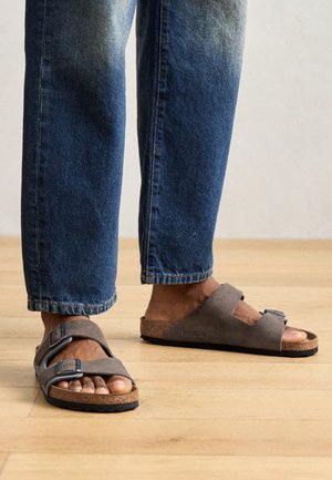 Graue Wildledersandalen mit zwei verstellbaren Riemen, Korkfußbett und schwarzer Gummisohle, getragen mit hellblauen, ausgebleichten Jeans in einer hellen Holzumgebung.