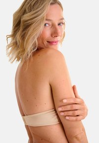 Donna bionda con capelli corti guarda oltre la spalla, indossando un reggiseno senza spalline beige, mentre tiene delicatamente il braccio contro uno sfondo neutro.