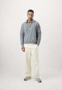 Ανοιχτό γκρι φλις πουλόβερ με φερμουάρ τριών τεράτων, που διαθέτει λευκό λογότυπο. Συνδυάζεται με κρεμ παντελόνι σε φαρδιά γραμμή και γκρι αθλητικά παπούτσια με μαύρες λεπτομέρειες.