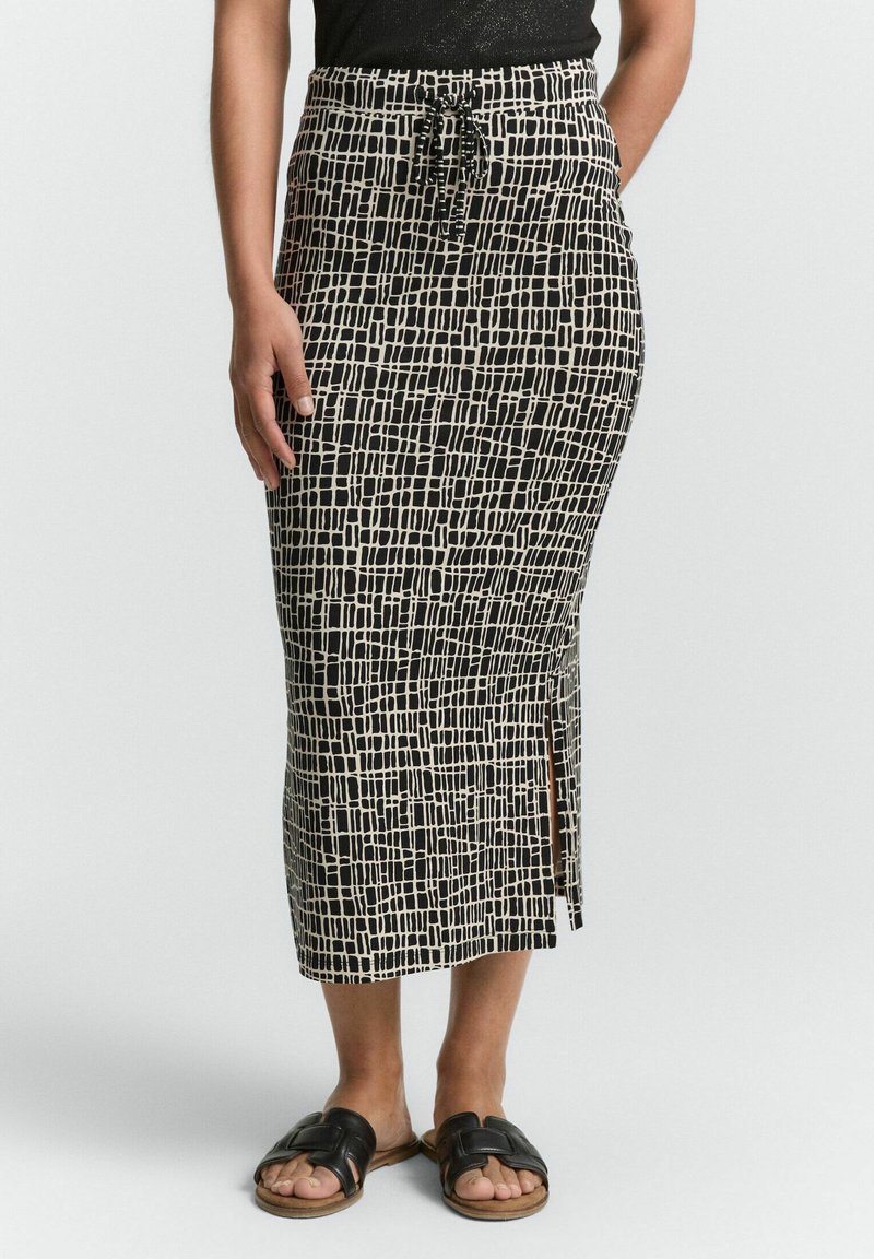 Jupe midi à motifs noirs et beiges avec taille à cordon, fente latérale et design légèrement ajusté. Accompagnée de sandales à glissière noires.