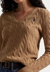 Femme portant un pull en maille torsadée beige à col en V avec un petit logo brodé de joueur de polo bleu marine, associé à un jean bleu et une ceinture noire.