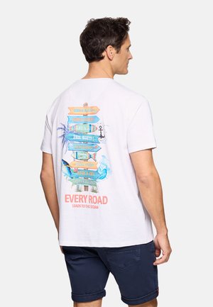 Hombre con camiseta blanca con un gráfico de poste de señalización temático del océano en colores y texto "Cada camino conduce al océano", lleva pantalones cortos oscuros, de perfil.