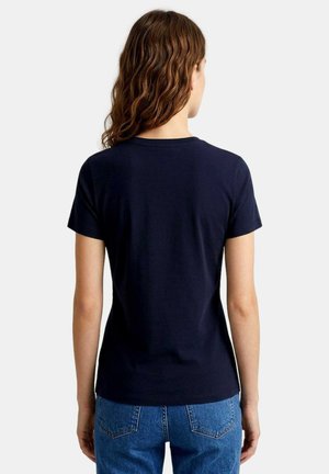 Donna con capelli castani mossi che indossa una semplice T-shirt blu navy a maniche corte e jeans blu, vista di spalle su sfondo bianco.