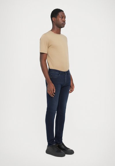 Tricou din bumbac bej și blugi skinny de culoare albastră închisă, purtați cu adidași negri, având un design simplu și texturi fine.