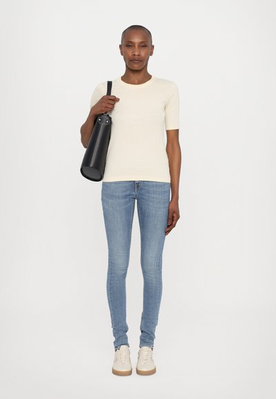 Camiseta corta de manga en beige claro, jeans ajustados azules y zapatillas de crema. Bolso negro de hombro con un acento de herraje circular.