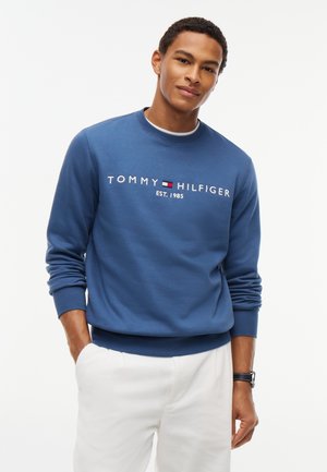 Homme portant un sweat-shirt bleu Tommy Hilfiger avec logo et un pantalon blanc, debout les mains dans les poches devant un fond blanc.