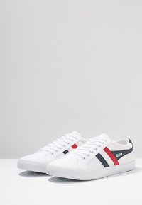 Sneaker in tela bianca con accenti a righe rosse e navy, suola in gomma testurizzata e logo del brand sul lato per un tocco di stile.