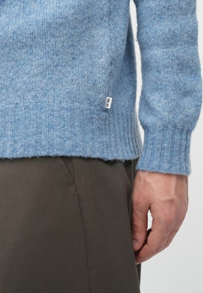 Gaiši zila vilnas džemperis ar ribinātām piedurknēm un apakšmalu, ar auduma struktūru un mazu zīmola etiķeti tuvu apakšējai šuvei.