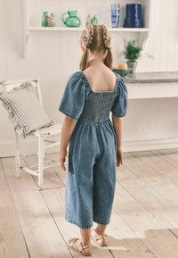 Denim jumpsuit med puffærmer, firkantet ryg og smocked body. Beskårne ben og afslappet pasform. Beige sandaler. Baggrund med lys trægulv.