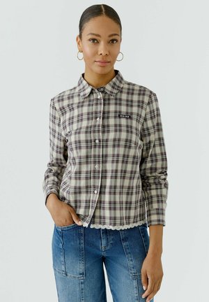 Frau mit zurückgebundenem Haar, trägt goldene Creolen, ein kariertes Hemd mit Knöpfen und dem "OUI OUI" Aufnäher sowie blaue Jeans mit großen Taschen.